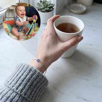 Thumbnail for Hand holding tea cup while wearing a Photo Projection Bracelet by Brother & Sisters, with a smiling baby in background.